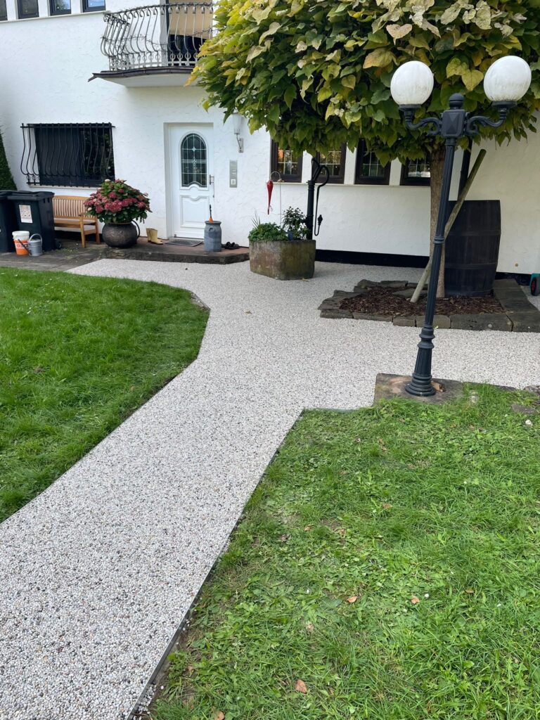 Gepflasterter Gartenweg vor einem weißen Wohnhaus mit Eingangstür, Fenstern und Balkon.